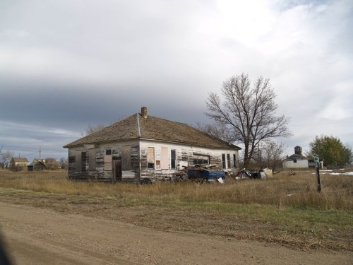 Abandoned house with peeling paint, surrounded by overgrown grass and scattered debris under a cloudy sky.