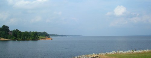 A serene lake view with calm waters, green trees along the shore, and a cloudy sky in the background.