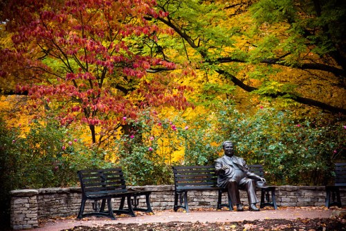 A bronze statue of a seated man surrounded by colorful autumn trees and benches in a serene park setting.