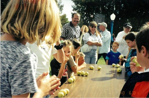 A group of children and adults participate in a fun outdoor event, with a table of apples in front of them.