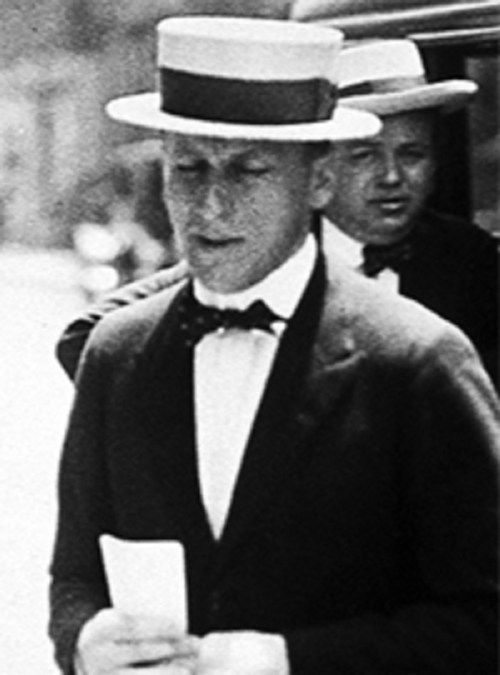 A man in a bow tie and straw hat holds a paper, with another man in a hat visible in the background.