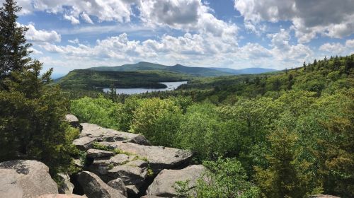 A scenic view of lush green hills and a lake under a partly cloudy sky, seen from a rocky overlook.