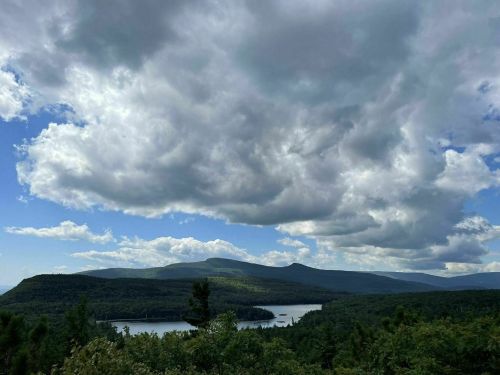 A scenic view of mountains and a lake under a cloudy sky, surrounded by lush greenery.