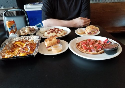 A table with various dishes: loaded fries, a sandwich, a small pizza, and dipping sauces, with a soda can nearby.