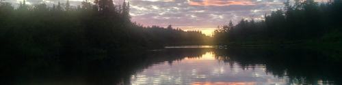 Sunset over a calm lake, surrounded by trees, with colorful clouds reflecting on the water's surface.