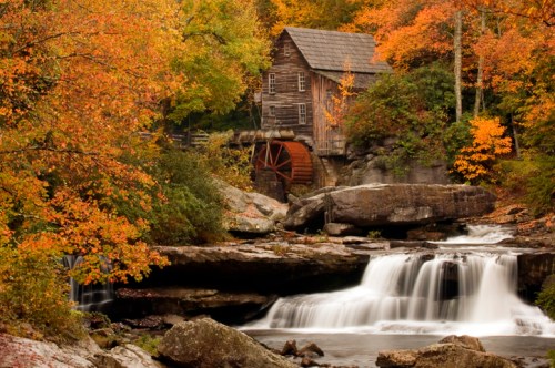 A rustic wooden mill beside a waterfall, surrounded by vibrant autumn foliage in shades of orange and yellow.