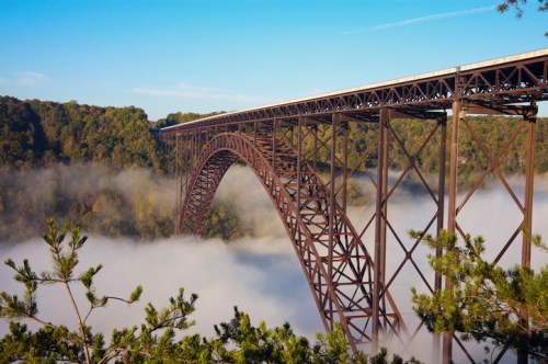 A large, arched bridge spans a foggy valley, surrounded by lush green trees and a clear blue sky.