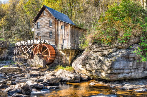 A rustic wooden mill with a water wheel beside a rocky stream, surrounded by trees in autumn colors.