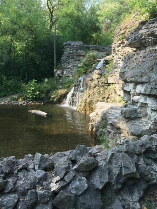 A serene waterfall cascades over rocky cliffs into a calm pool, surrounded by lush greenery.