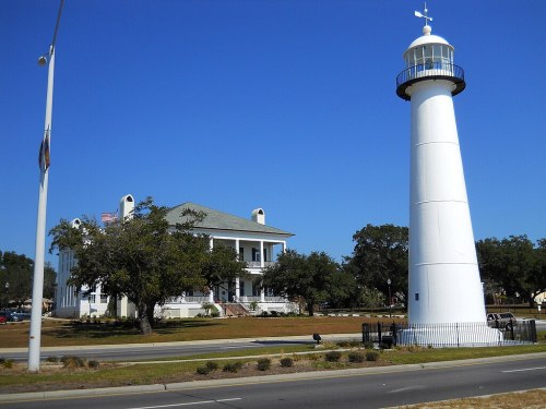 A tall white lighthouse beside a historic building, set against a clear blue sky and green trees.