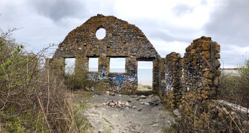 Ruins of a stone building with open windows, surrounded by overgrown vegetation and a cloudy sky in the background.