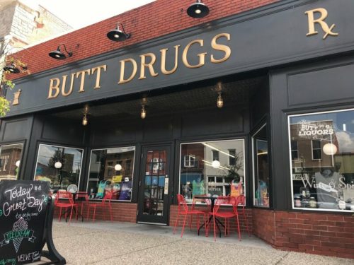 A storefront with the sign "BUTT DRUGS" and red outdoor seating, featuring a chalkboard sign promoting a special event.