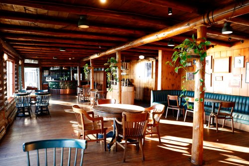 Cozy interior of a rustic restaurant with wooden beams, tables, and plants, featuring a bar in the background.