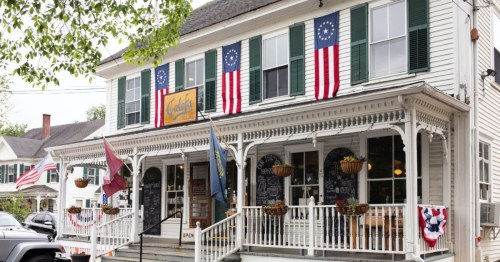 A charming white building with green shutters, featuring flags and a welcoming porch, labeled "Café."