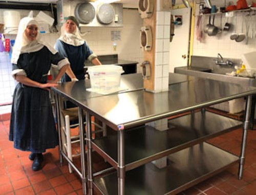 Two women in traditional attire stand in a kitchen with stainless steel equipment and a large work table.