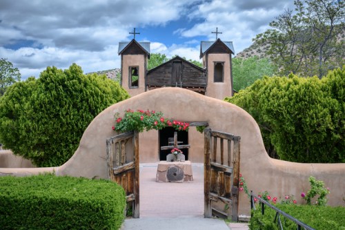 A rustic adobe building with crosses, surrounded by greenery and flowers, under a cloudy sky.