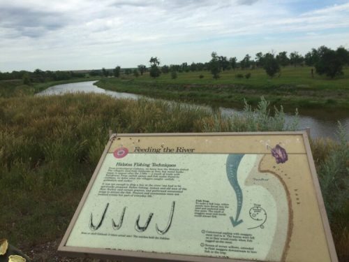 A sign about fishing techniques beside a river, with green fields and trees in the background under a cloudy sky.