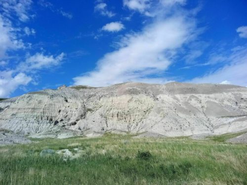 A rugged hillside with layered rock formations under a blue sky with scattered clouds, surrounded by green grass.