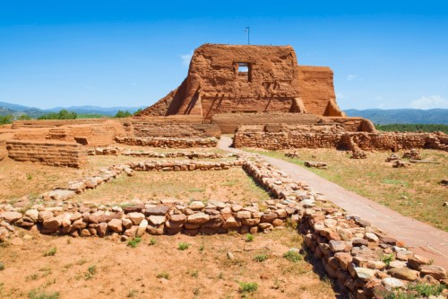Ancient adobe ruins with a stone pathway and blue sky in the background, surrounded by arid landscape.