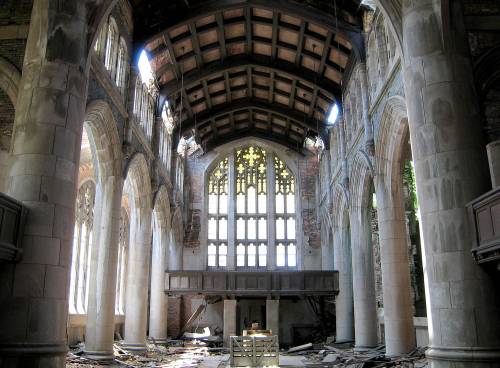 Interior of an abandoned church, featuring tall arches, stained glass windows, and a wooden ceiling, with debris on the floor.
