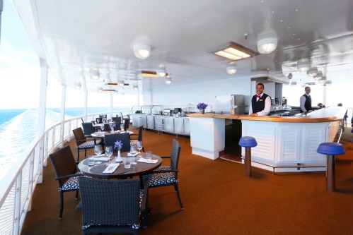A spacious dining area on a ship with tables, a bar, and a view of the ocean, featuring a smiling staff member.