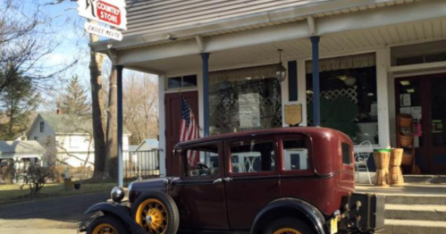 A vintage maroon car parked in front of a quaint country store with a sign and American flag.