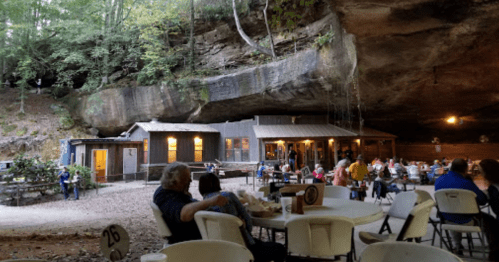 A rustic restaurant under a large rock overhang, with diners enjoying meals in a natural cave setting.