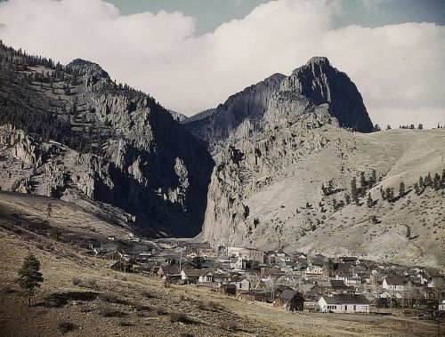 A scenic view of a small town nestled in a valley surrounded by rugged mountains and cloudy skies.