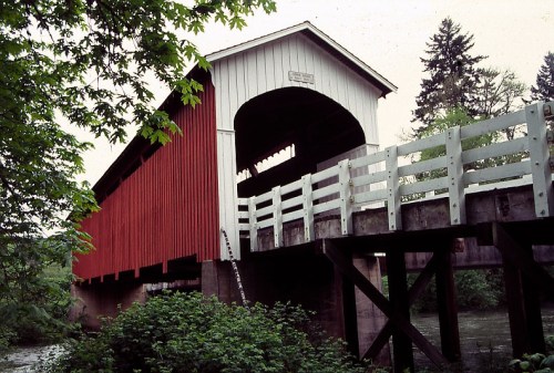 A red covered bridge spans a river, surrounded by lush greenery and trees under a cloudy sky.