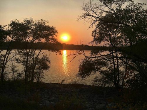 Sunset over a calm lake, with silhouettes of trees framing the scene and a warm orange glow reflecting on the water.