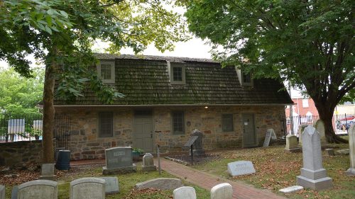Historic stone building with a sloped roof, surrounded by trees and gravestones in a cemetery setting.