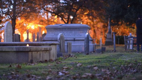 A serene cemetery scene at dusk, featuring gravestones and a softly lit mausoleum surrounded by autumn leaves.