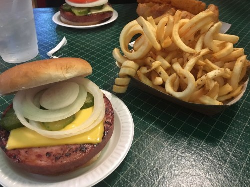 A plate with a sandwich featuring a grilled patty, cheese, pickles, and onions, alongside a serving of curly fries.