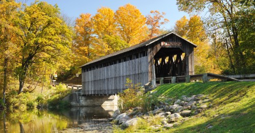 A wooden covered bridge spans a calm stream, surrounded by vibrant autumn foliage and a clear blue sky.