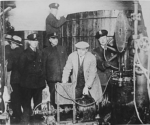 Historical black-and-white photo of men in suits and police uniforms around machinery in a factory setting.