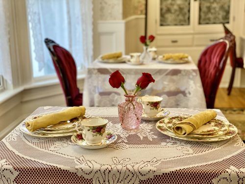 A cozy dining setup with floral china, rolled crepes, and a vase of red roses on a lace tablecloth.