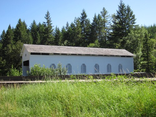A white covered bridge surrounded by tall green grass and trees under a clear blue sky.