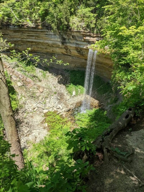 A serene waterfall cascades into a rocky basin, surrounded by lush greenery and steep, layered cliffs.