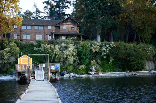 A wooden dock leads to a charming house on a hillside, surrounded by trees and greenery by the water's edge.