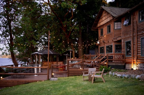 A cozy wooden lodge by the water, surrounded by trees and string lights, with a deck and seating area.