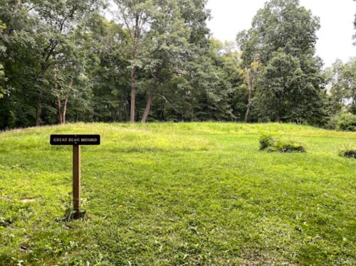 A grassy area with a sign reading "Great Bear Mound" surrounded by trees under a cloudy sky.