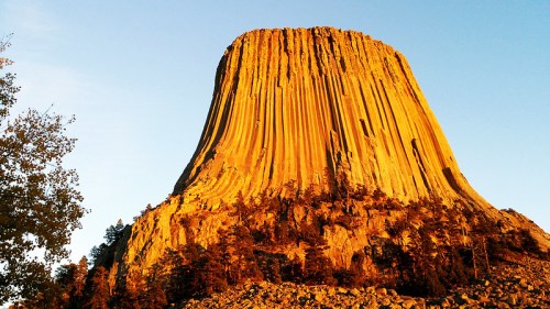 A towering rock formation with vertical columns, illuminated by golden sunlight, surrounded by trees and a clear sky.
