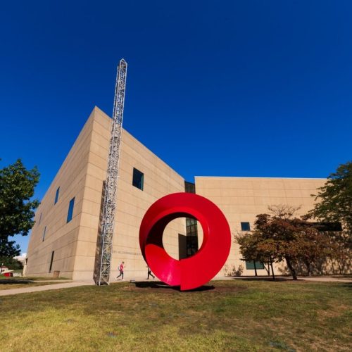 A large red circular sculpture stands in front of a modern building under a clear blue sky.
