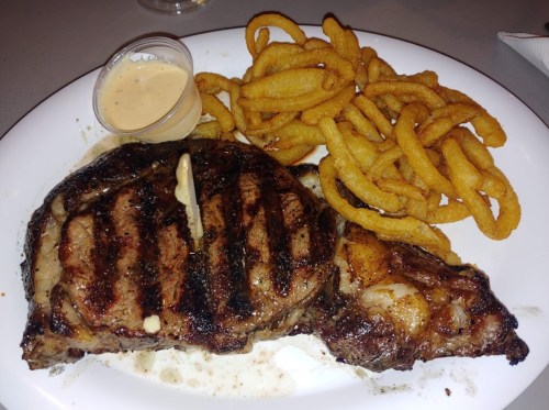 A grilled steak with grill marks, served with curly fries and a small cup of dipping sauce on a white plate.