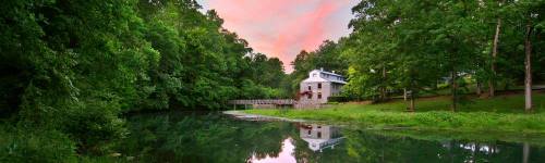 A serene river scene with a house and trees, reflecting a colorful sunset in the water.