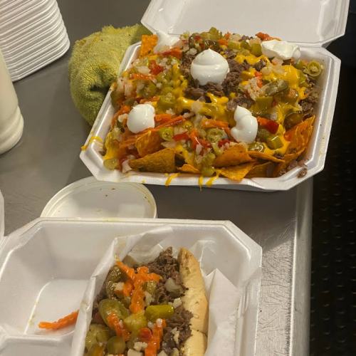 A takeout container with loaded nachos topped with cheese, sour cream, and jalapeños, alongside a hot dog with toppings.
