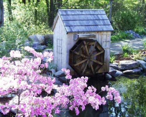 A wooden watermill beside a pond, surrounded by blooming pink flowers and lush greenery.