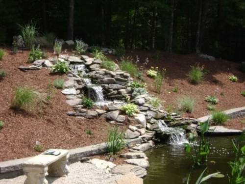 A serene garden with a stone waterfall cascading into a pond, surrounded by lush greenery and mulch.