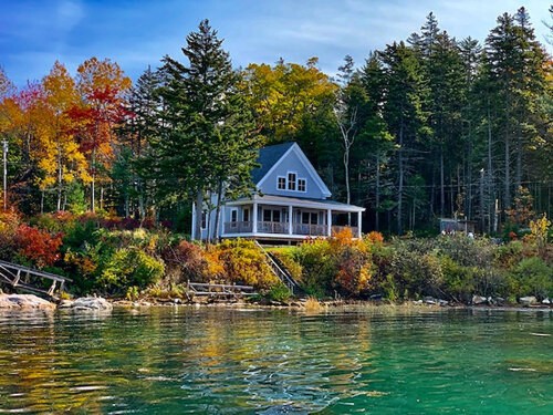 A serene lakeside house surrounded by colorful autumn trees and calm water, reflecting the vibrant fall foliage.
