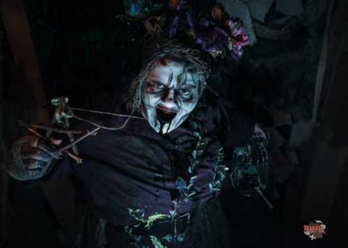 A creepy figure with dark makeup and flowers in their hair, holding a puppet and appearing menacing in a dimly lit setting.